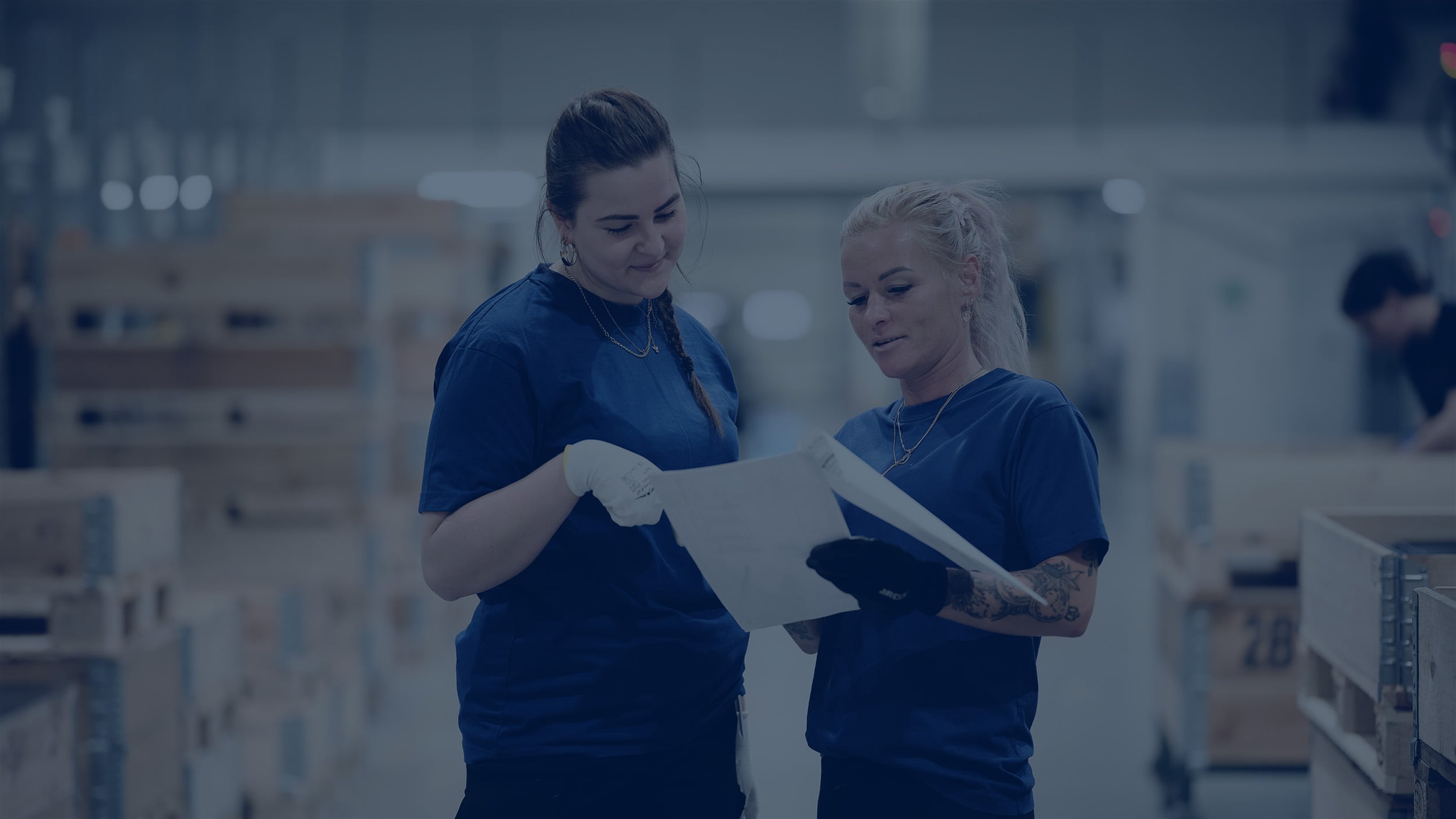 Zwei Fabrikarbeiter in blauen Uniformen überprüfen ein Dokument in einer industriellen Umgebung.