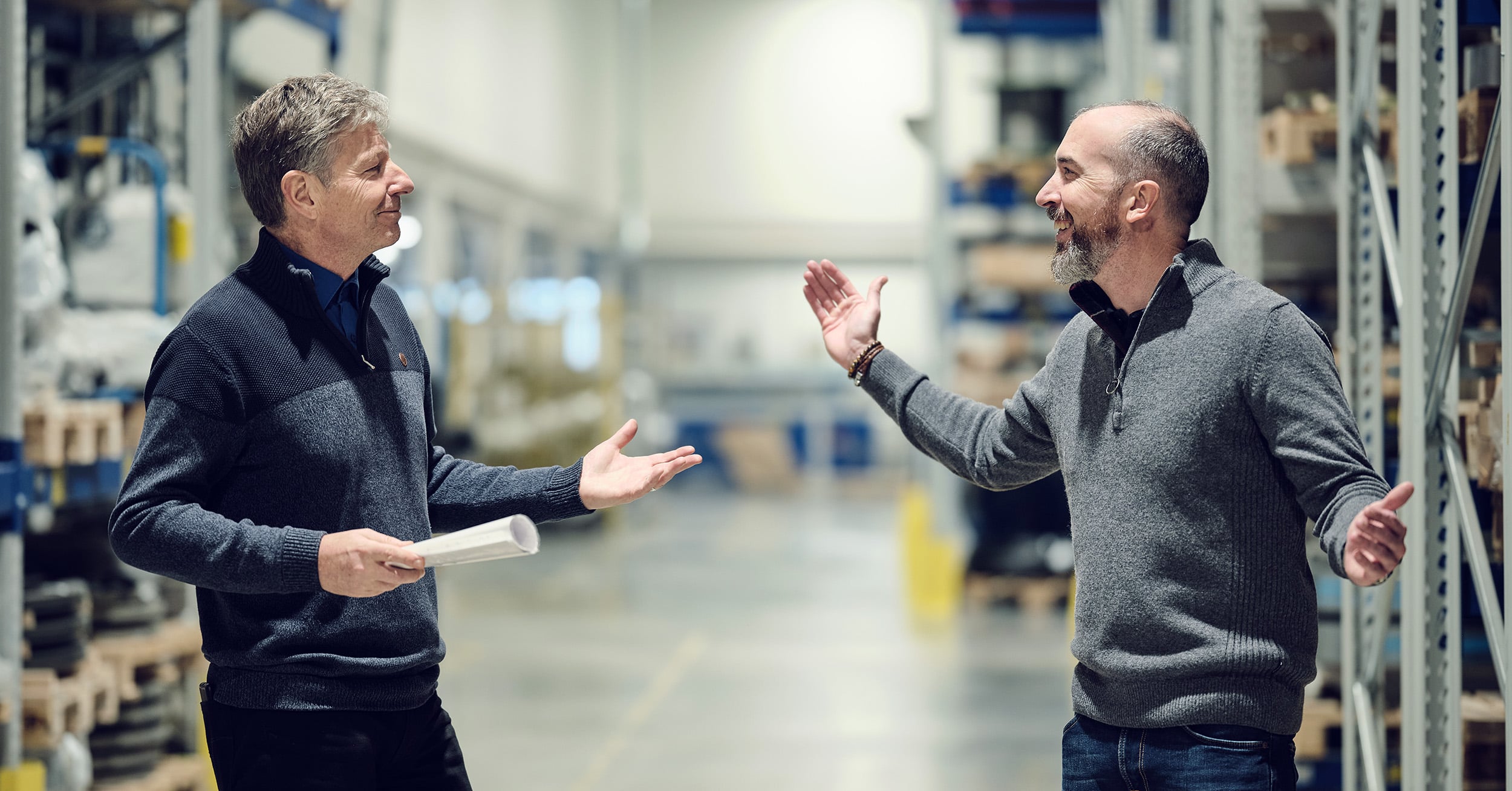 Twee mannen hebben een gesprek in een goed verlichte industriële opslagruimte, een houdt een document vast.