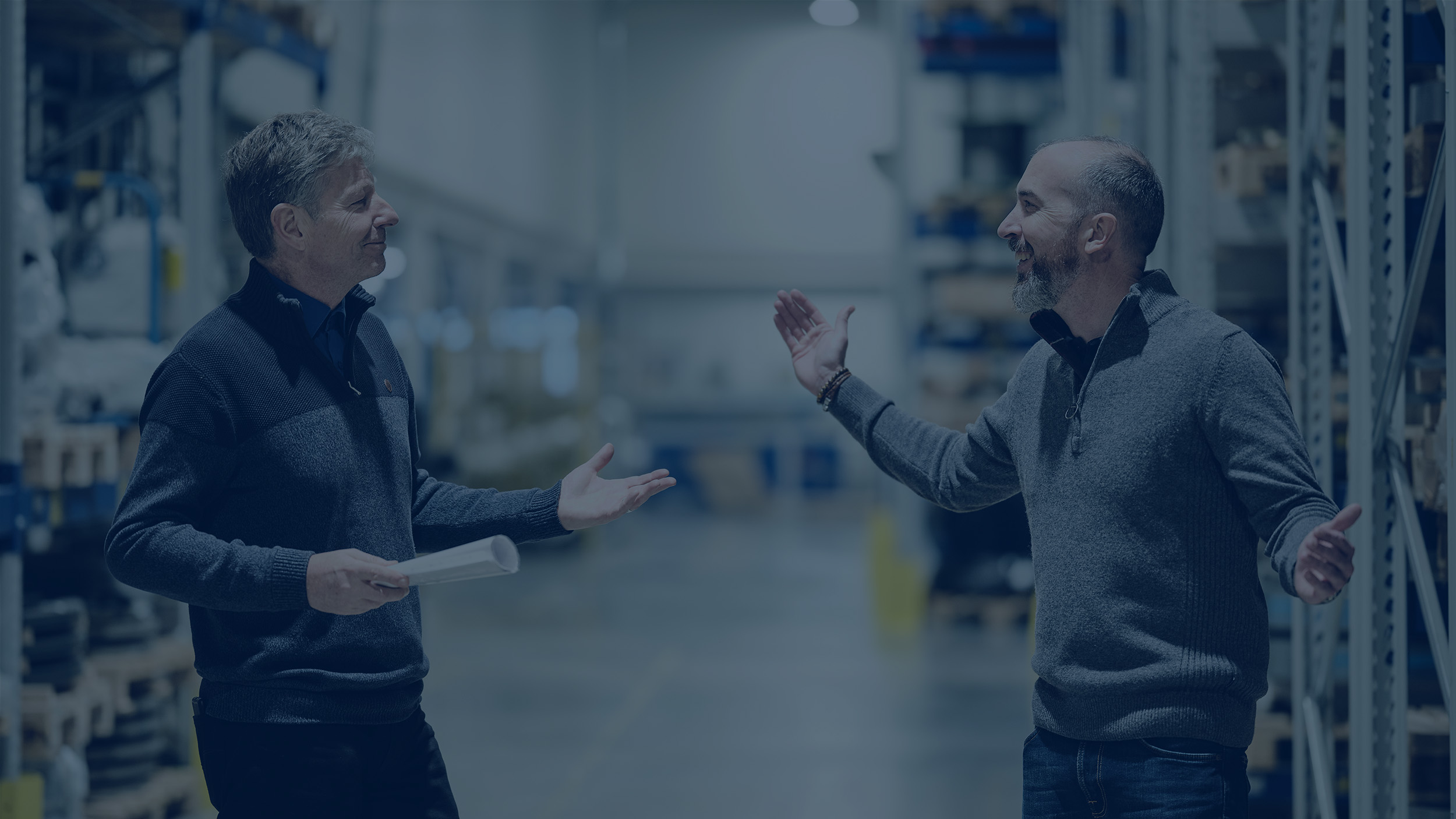 Due uomini in un magazzino industriale impegnati in una conversazione amichevole, discutendo un documento.